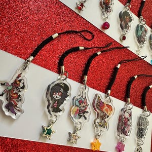 May include: A collection of clear acrylic charms featuring colorful cartoon characters, each attached to a black lanyard with a silver clasp. Some charms have star-shaped or round dangling accents. The background is red glitter.