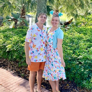 May include: A couple wearing matching shirts and a dress with a colorful balloon and Mickey Mouse print. The man is wearing orange shorts and sandals. The woman is wearing a light blue cardigan and yellow sandals. They are standing on a brick pathway in front of green bushes.