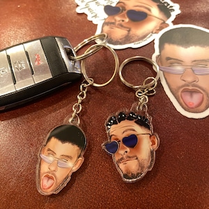 May include: Two clear acrylic keychains featuring cartoon portraits of a man with black hair and a beard wearing sunglasses. One keychain has the man wearing heart-shaped sunglasses and the other has the man wearing rectangular sunglasses. Both men have their tongues sticking out.
