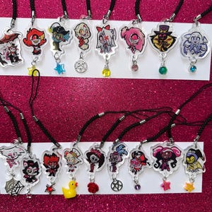 May include: A collection of clear acrylic charm necklaces featuring colorful cartoon characters. Each charm is attached to a black cord and includes a decorative charm, such as a star or a gem. The background is a sparkly pink.