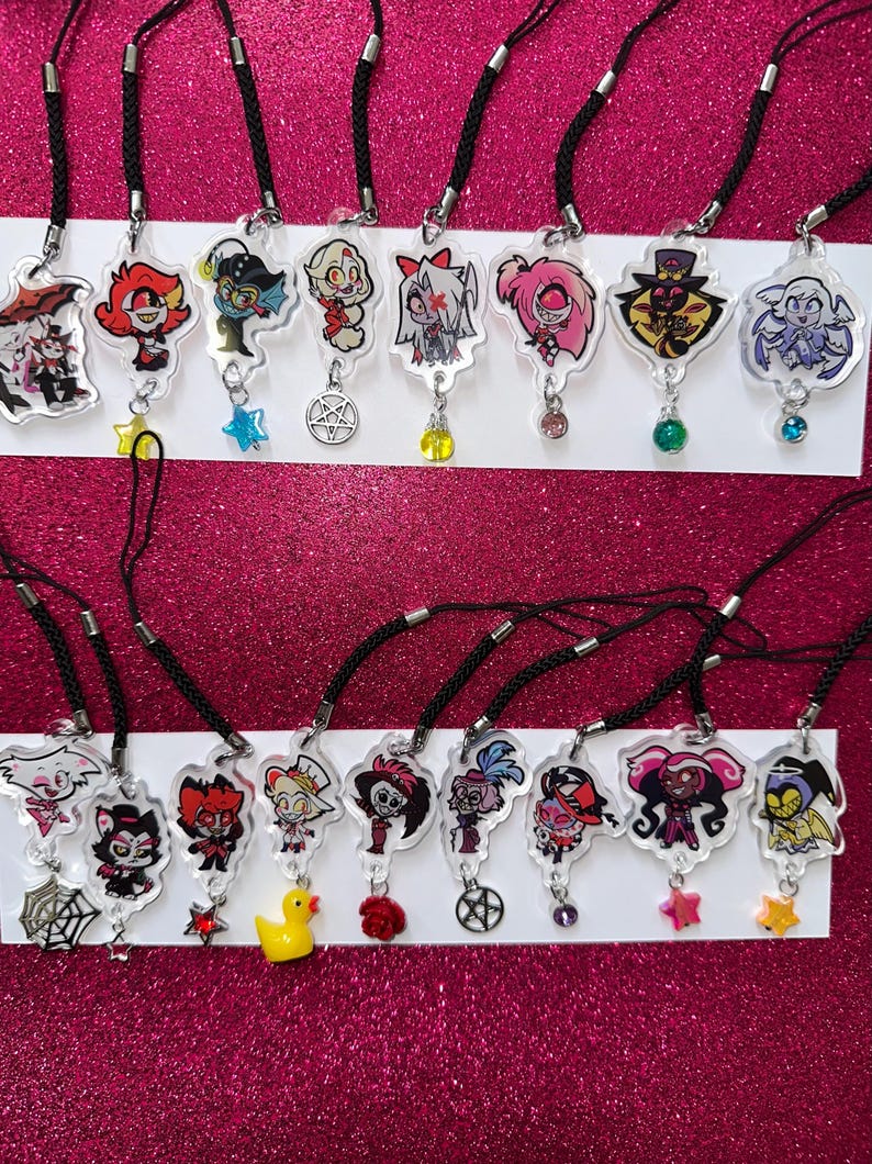 May include: A collection of clear acrylic charm necklaces featuring colorful cartoon characters. Each charm is attached to a black cord and includes a decorative charm, such as a star or a gem. The background is a sparkly pink.