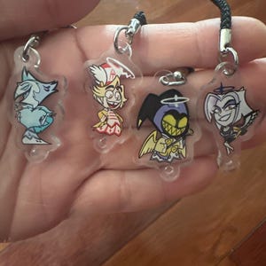May include: Four clear acrylic charms with cartoon character designs. Each charm has a silver-colored clasp and a black lanyard. The characters include a blue wolf, a blonde angel, a purple demon, and a white-haired character.