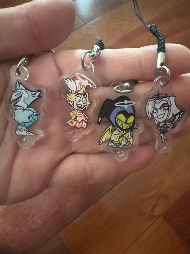 May include: Four clear acrylic charms with cartoon character designs. Each charm has a silver-colored clasp and a black lanyard. The characters include a blue wolf, a blonde angel, a purple demon, and a white-haired character.