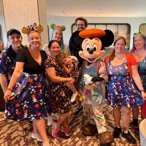 Pode incluir: Um grupo de pessoas vestindo roupas com tema Disney, incluindo vestidos e camisas com estampas de Halloween de ab&oacute;boras e fantasmas, ao lado de um personagem do Mickey Mouse vestido com um avental manchado de tinta e uma boina laranja.
