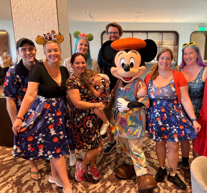 Pode incluir: Um grupo de pessoas vestindo roupas com tema Disney, incluindo vestidos e camisas com estampas de Halloween de ab&oacute;boras e fantasmas, ao lado de um personagem do Mickey Mouse vestido com um avental manchado de tinta e uma boina laranja.