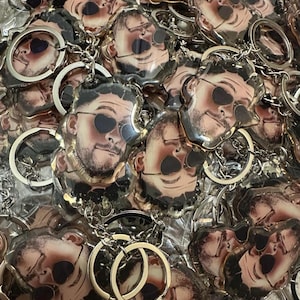 Op de afbeelding: Een verzameling sleutelhangers met een portret van een persoon met een zonnebril. Elke sleutelhanger heeft een zilverkleurige ketting en een metalen ring. De afbeelding op de sleutelhanger is een close-up van het gezicht van een persoon.