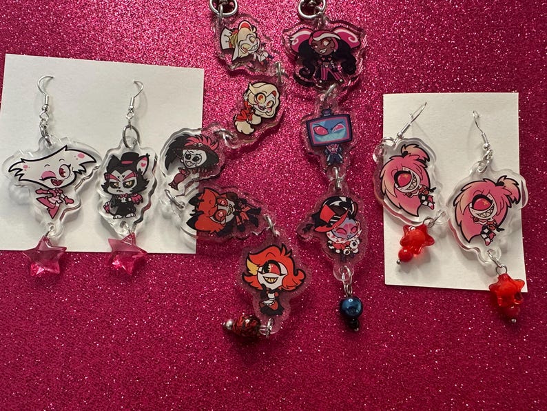 K&ouml;nnte beinhalten: Eine Sammlung von Acryl-Ohrringen und Schl&uuml;sselanh&auml;ngern mit Cartoon-Charakteren. Die Ohrringe haben silberne Haken und sternf&ouml;rmige oder perlenf&ouml;rmige Akzente. Die Schl&uuml;sselanh&auml;nger sind durch silberne Ketten verbunden und haben verschiedene Charakterdesigns.