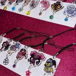 May include: Collection of clear acrylic charms featuring colorful cartoon characters, each with a black cord and a decorative charm. Charms include star, pentagram, and gem accents. The background is a sparkly pink surface.