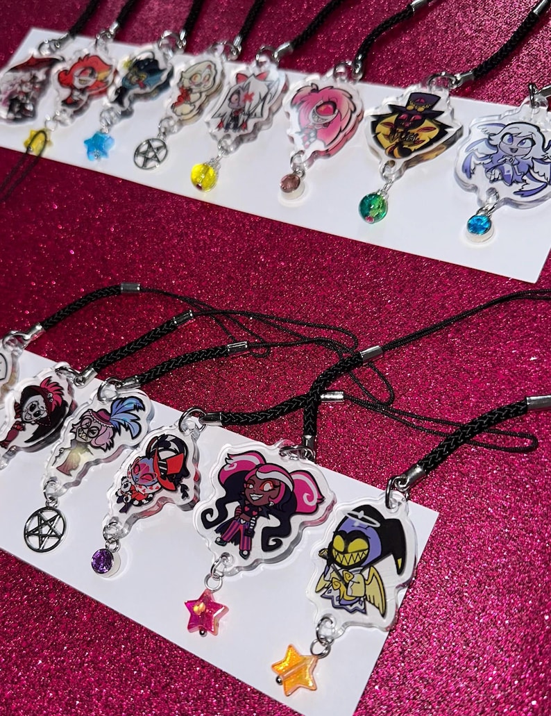 May include: Collection of clear acrylic charms featuring colorful cartoon characters, each with a black cord and a decorative charm. Charms include star, pentagram, and gem accents. The background is a sparkly pink surface.