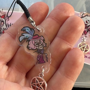 May include: Clear acrylic charm featuring a cartoon character with pink hair, a hat, and a cane. The charm hangs from a black cord and a silver-colored pentagram charm. Additional charms are visible in the background.