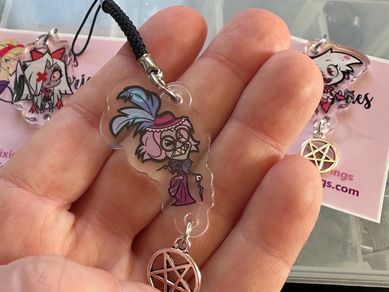 May include: Clear acrylic charm featuring a cartoon character with pink hair, a hat, and a cane. The charm hangs from a black cord and a silver-colored pentagram charm. Additional charms are visible in the background.