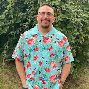 May include: A man wearing a light blue short-sleeved shirt with a floral and polka dot pattern. The shirt features a repeating pattern of red roses, blue flowers, and white polka dots. Mickey Mouse heads are also part of the pattern.