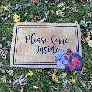 May include: Rectangular sign with the words "Please Come Inside" in black script. A cartoon Deadpool character is on the right, holding a paper that says "That's what she said!" The sign has a brown background and a black border.