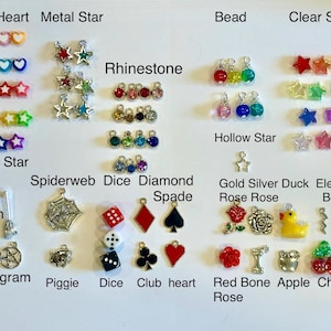 May include: A collection of small charms in various shapes and colors, including hearts, stars, dice, and roses. The charms are arranged on a white surface and labeled with their names, such as "Solid Heart" and "Metal Star".