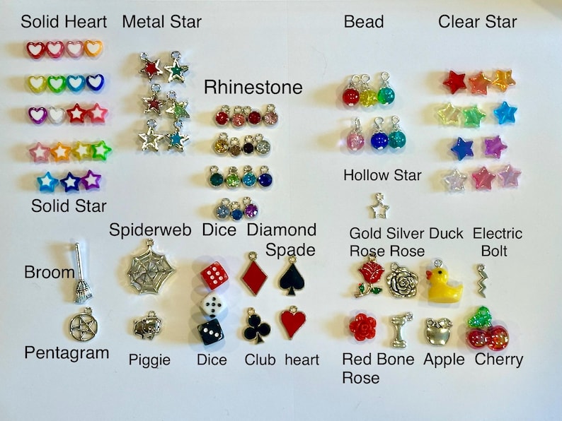 May include: A collection of small charms in various shapes and colors, including hearts, stars, dice, and roses. The charms are arranged on a white surface and labeled with their names, such as "Solid Heart" and "Metal Star".