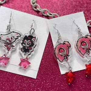 K&ouml;nnte beinhalten: Transparente Acryl-Ohrringe mit Comicfiguren mit rosa und roten Akzenten. Die Ohrringe haben silberne Haken und stern- oder edelsteinf&ouml;rmige Anh&auml;nger. Sie werden auf wei&szlig;en Karten vor einem pinkfarbenen Glitzerhintergrund pr&auml;sentiert.