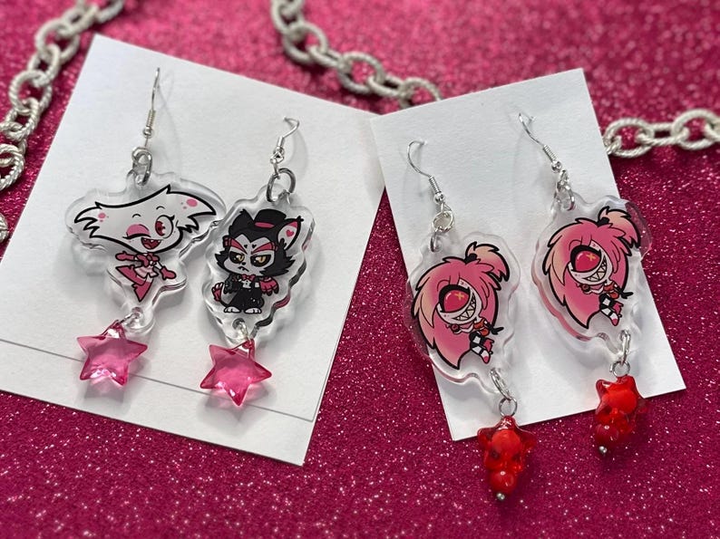 K&ouml;nnte beinhalten: Transparente Acryl-Ohrringe mit Comicfiguren mit rosa und roten Akzenten. Die Ohrringe haben silberne Haken und stern- oder edelsteinf&ouml;rmige Anh&auml;nger. Sie werden auf wei&szlig;en Karten vor einem pinkfarbenen Glitzerhintergrund pr&auml;sentiert.