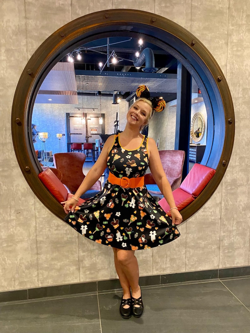Pode incluir: Um vestido preto sem mangas com um padr&atilde;o repetitivo de doces coloridos e &iacute;cones em forma de rato. O vestido tem um decote redondo, uma saia rodada e &eacute; usado com um cinto largo de cor laranja s&oacute;lida e uma tiara de orelhas a combinar.