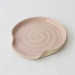 May include: A ceramic spoon rest in a soft pink hue with a light beige rim. The spoon rest has a circular shape with a slight indentation on one side. The centre of the rest features a spiral design.