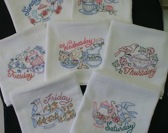 May include: Set of seven white tea towels with embroidered designs and days of the week. Each towel features a different bird, teapot, and floral design. The days of the week are embroidered in cursive script.