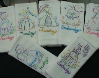 May include: Set of seven white tea towels with embroidered images of girls holding umbrellas and flowers. Each towel features a different day of the week, spelled out in colorful embroidery. The towels are perfect for adding a touch of vintage charm to your kitchen.