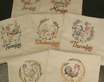 May include: Set of seven embroidered tea towels featuring a rooster and wheat design with each day of the week labeled. The towels are white with brown, green, and red embroidery.