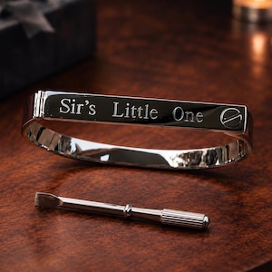 May include: A polished silver bracelet engraved with the words "Sir's Little One" rests on a wooden surface. A small tool is positioned nearby. A black gift box with a bow is visible in the background.