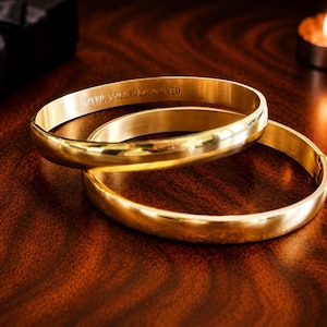 Peut inclure: Deux bracelets dorés sont posés sur une surface en bois sombre. Le bracelet supérieur porte un texte gravé. Un coffret cadeau noir avec un nœud est en arrière-plan, ainsi qu'une bougie allumée. Les bracelets sont de couleur or chaud et brillant.