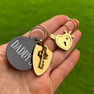 May include: A gold-toned heart-shaped padlock pendant with a small crown and a red accent on a chain, paired with a circular black tag engraved with the word DADDY'S and a gold-toned shield-shaped charm featuring a cutout key design on a ring.