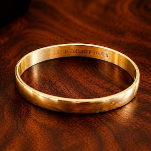 May include: A gold-colored bangle bracelet with the engraved words "LOVED, COLLARED, OWNED" is displayed on a dark wooden surface. A black gift box and a lit candle are in the background, creating a warm ambiance. The bracelet is a simple, elegant design.