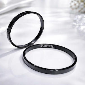 Puede incluir: Dos pulseras de metal negro. Una pulsera está grabada con la palabra "Owned", y la otra con la palabra "Protected". Las pulseras son circulares y tienen un cierre de broche.