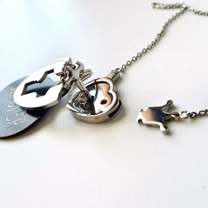 May include: A silver-toned metal necklace featuring a heart-shaped pendant with a small red stone, a key charm, and a circular tag with the word SISTER engraved. The set includes a separate puzzle-piece shaped charm on a matching silver chain.
