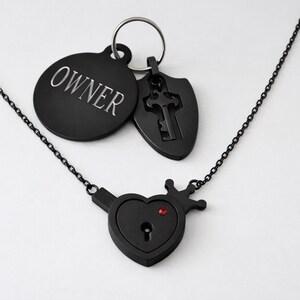 May include: A black metal necklace featuring a heart-shaped pendant with a keyhole and a small red stone. Attached to a silver-toned ring are two black charms: a circular tag engraved with the word OWNER and a shield-shaped charm with a key cutout.