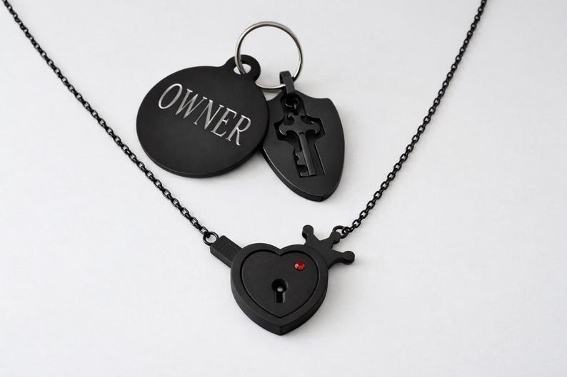 May include: A black metal necklace featuring a heart-shaped pendant with a keyhole and a small red stone. Attached to a silver-toned ring are two black charms: a circular tag engraved with the word OWNER and a shield-shaped charm with a key cutout.