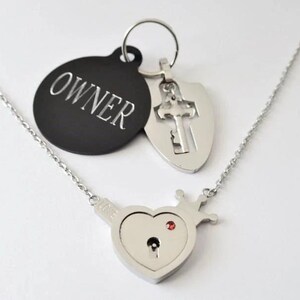 May include: A silver-toned necklace featuring a heart-shaped pendant with a crown detail, a keyhole, and a small red stone. Attached are two charms: a black circular tag engraved with the word OWNER and a silver shield-shaped charm with a cross cutout.