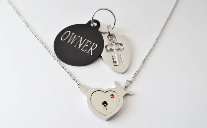 May include: A silver-toned necklace featuring a heart-shaped pendant with a crown detail, a keyhole, and a small red stone. Attached are two charms: a black circular tag engraved with the word OWNER and a silver shield-shaped charm with a cross cutout.