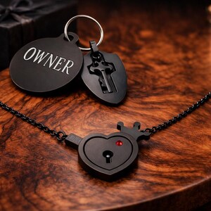 May include: A black metal necklace featuring a heart-shaped pendant with a small crown and a red stone, connected to a chain. A circular tag engraved with the word OWNER and a shield-shaped charm with a key cutout are attached to a metal ring.
