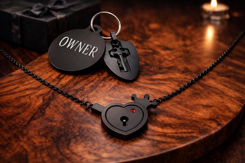 May include: A black metal necklace featuring a heart-shaped pendant with a small crown and a red stone, connected to a chain. A circular tag engraved with the word OWNER and a shield-shaped charm with a key cutout are attached to a metal ring.