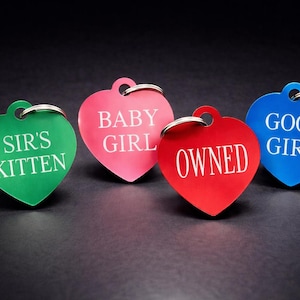 May include: Four heart-shaped pet tags in green, pink, red, and blue. Each tag has a silver ring and features white text: "SIR'S KITTEN", "BABY GIRL", "OWNED", and "GOOD GIRL". The tags are on a dark surface.
