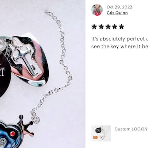 May include: A silver-toned chain necklace featuring a round black pendant engraved with the text THE OWNER OF MY HEART and 10/31/22. Attached are a silver key-shaped charm and a heart-shaped charm with a crown detail and a small red accent.