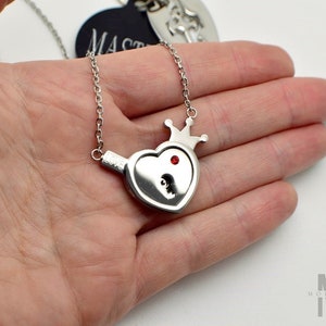 May include: A silver-toned heart-shaped pendant necklace featuring a small crown at the top, a single red crystal accent, and a keyhole cutout. The word LOVE is engraved on the side of the pendant, which hangs from a delicate silver chain.