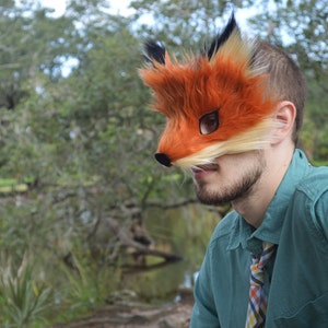 Op de afbeelding: Een persoon draagt ​​een bruin en oranje vossenmasker met zwarte vachtdetails. Het masker bedekt de ogen en neus van de persoon. De persoon draagt ​​een tealgroen shirt met een veelkleurige gepatenteerde stropdas.