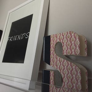 May include: A white framed picture with the word "FRIENDS" in black text on a black background. The picture is hanging on a wall next to a decorative letter "S" made of fabric with a red and white floral pattern.