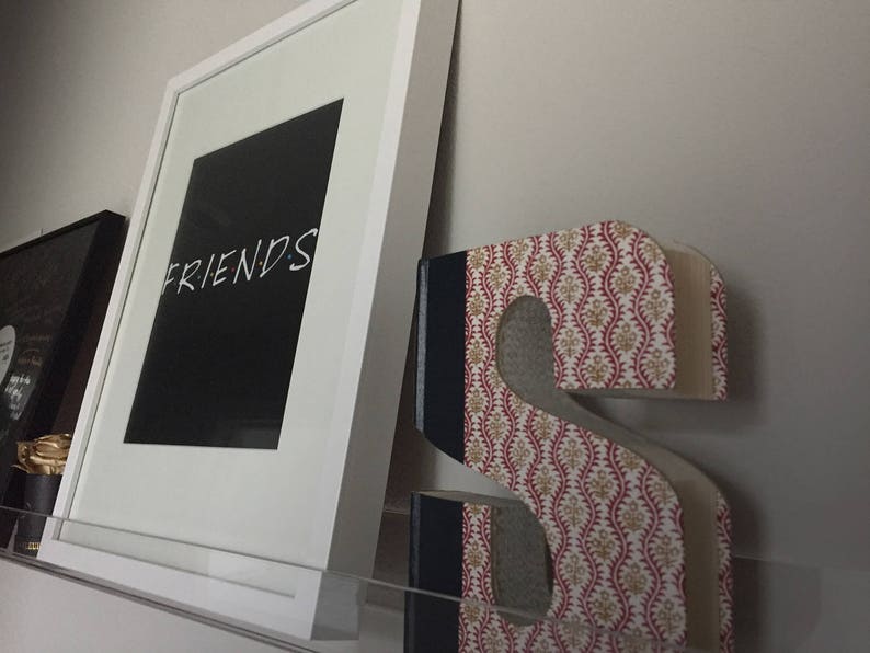 May include: A white framed picture with the word "FRIENDS" in black text on a black background. The picture is hanging on a wall next to a decorative letter "S" made of fabric with a red and white floral pattern.