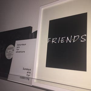 May include: A white framed print with a black background and the word "FRIENDS" in white with colorful dots. The print is hanging on a white wall next to two other prints with black backgrounds and white text.