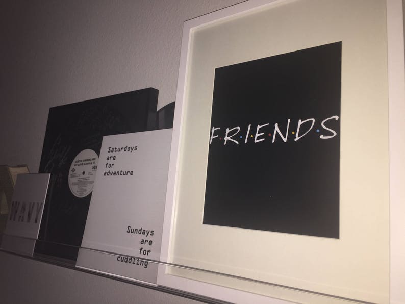 May include: A white framed print with a black background and the word "FRIENDS" in white with colorful dots. The print is hanging on a white wall next to two other prints with black backgrounds and white text.