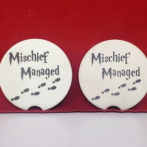 May include: Two white ceramic coasters with the text "Mischief Managed" in black lettering and a graphic of footprints.