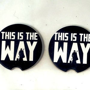 May include: Two black round coasters with a white silhouette of a figure with a staff and a small creature. The text "This is the Way" is printed in white on the coasters.