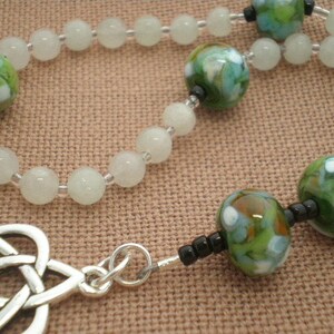 May include: A white and green beaded necklace with a silver Celtic knot pendant. The beads are made of glass and have a speckled pattern.