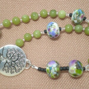 Puede incluir: Un collar de cuentas verdes y plateadas con un dije de árbol de la vida. Las cuentas son verdes y blancas con diseños florales.